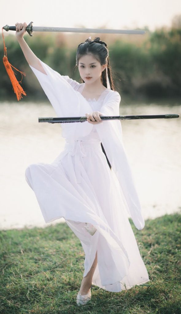 Asian woman practicing Tai Chi sword kata in a white dress at a serene park setting.